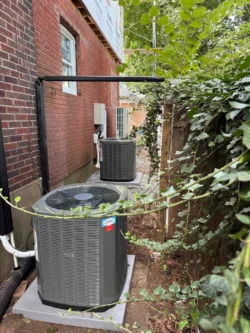 Two outdoor air conditioning units sit on concrete pads next to a brick building, surrounded by greenery and a wooden fence.