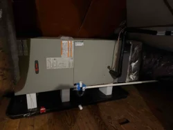 A central HVAC air handler unit is installed in an attic on a wooden floor, with pipes and tubing connected for drainage and operation.